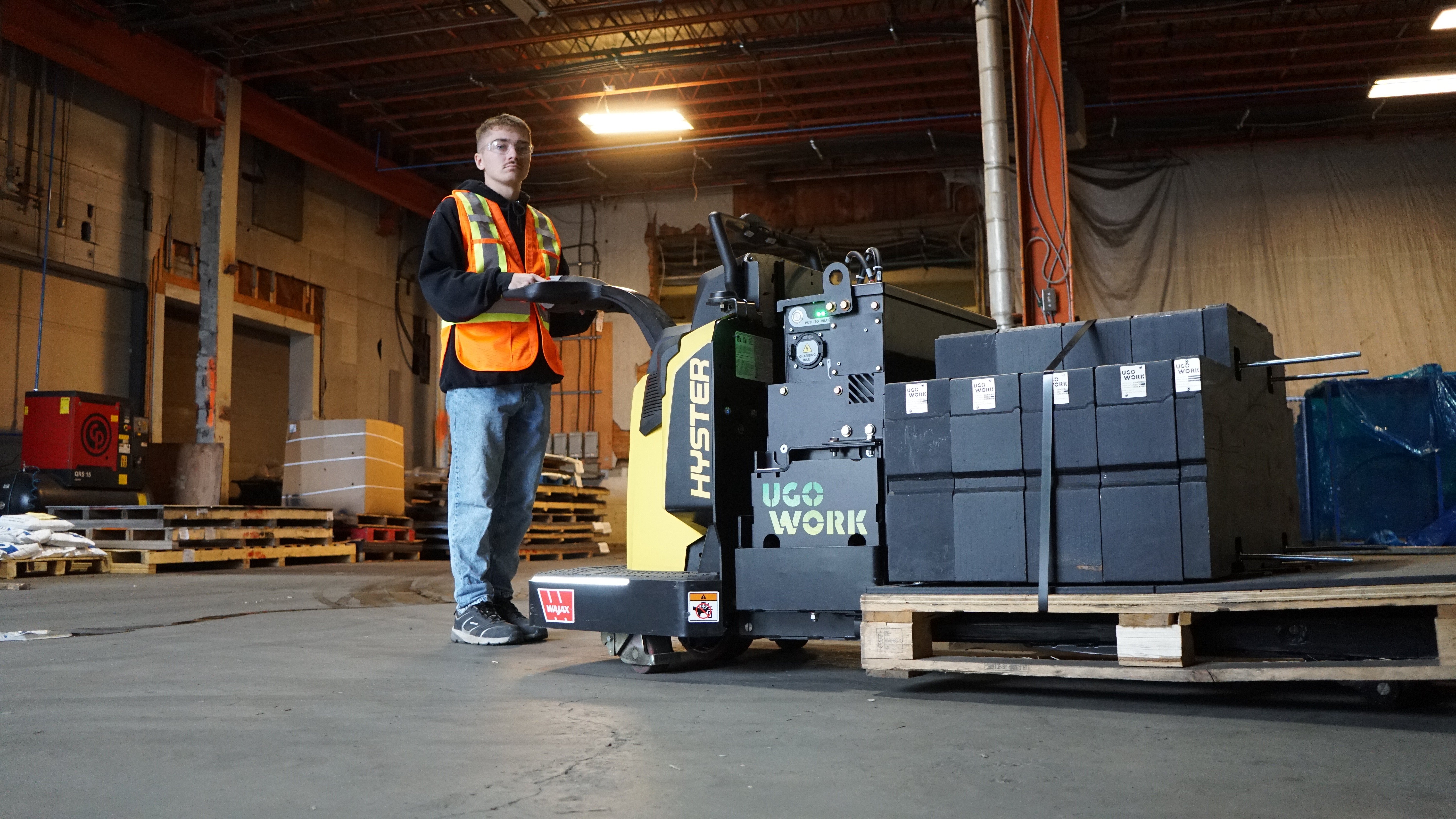 A person wearing a safety vest operates a Hyster pallet jack powered by a UgoWork lithium-ion battery to move boxes in a large warehouse with stacked pallets and industrial equipment.
